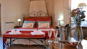Una cama bien hecha con colcha roja y blanca, dos almohadas y un dosel. Hay una mesa con dos copas de champán y un jarrón con flores.