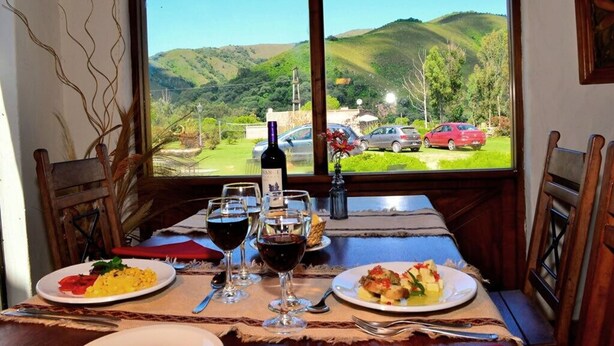 Una mesa de comedor con platos de comida y copas de vino, una botella de vino, y una vista de un paisaje verde con colinas y coches al fondo.