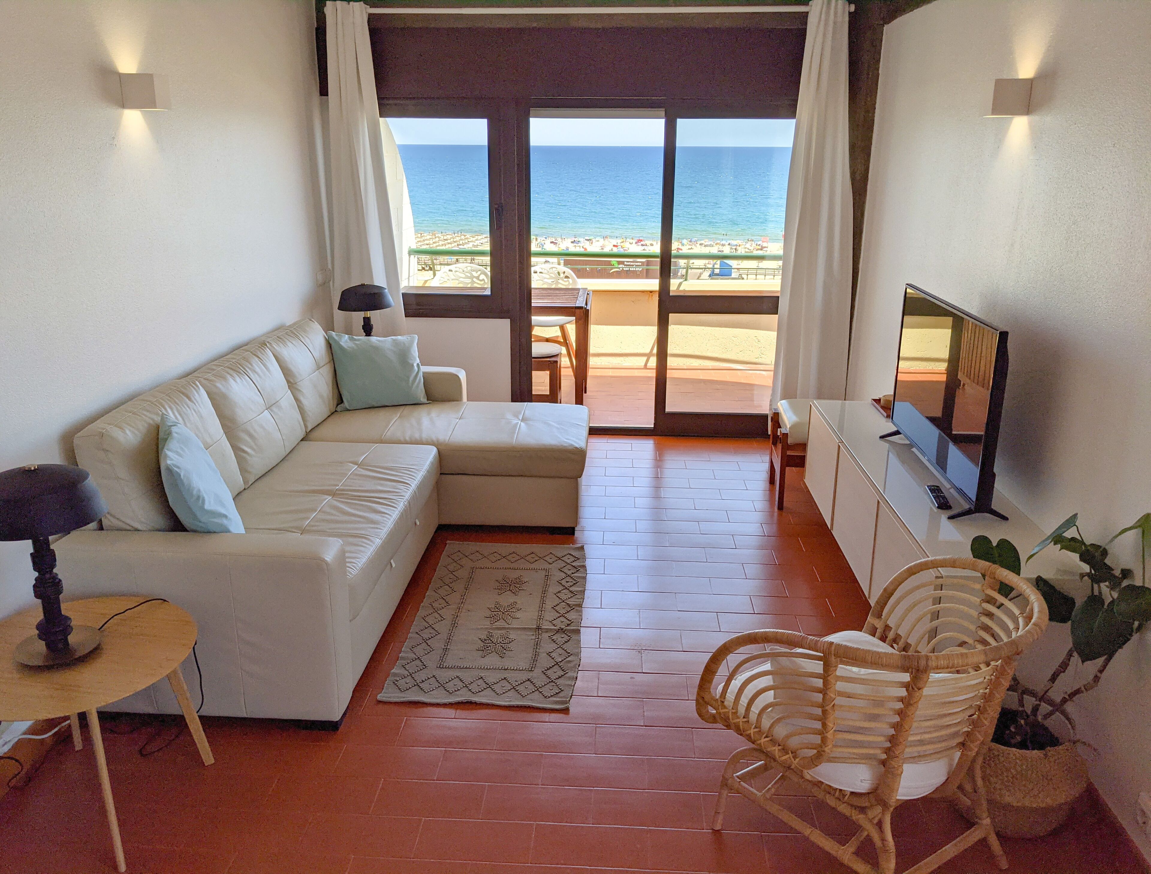 A living room with a sofa, a TV, a small table, and a wicker chair. There is a view of the beach through the glass door.