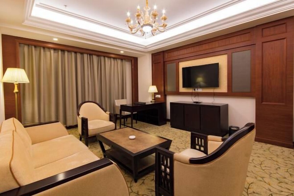 A hotel room with a sofa, armchair, coffee table, TV, and a desk with a lamp.