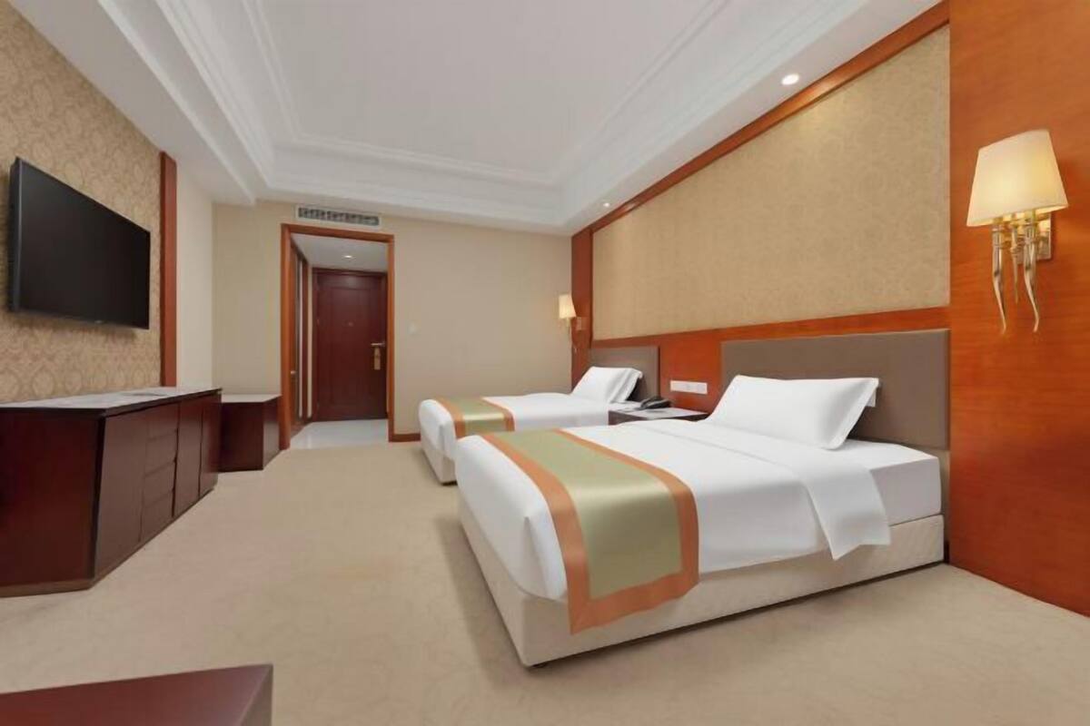 A hotel room with two beds, a desk, a TV, and a wall-mounted lamp.