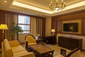 Quarto de hotel com sofá, poltrona, mesa de centro, televisão, escrivaninha e uma janela grande.