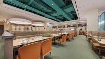 A modern restaurant with booths and tables, a high ceiling with green lighting, and a view of the city through large windows.