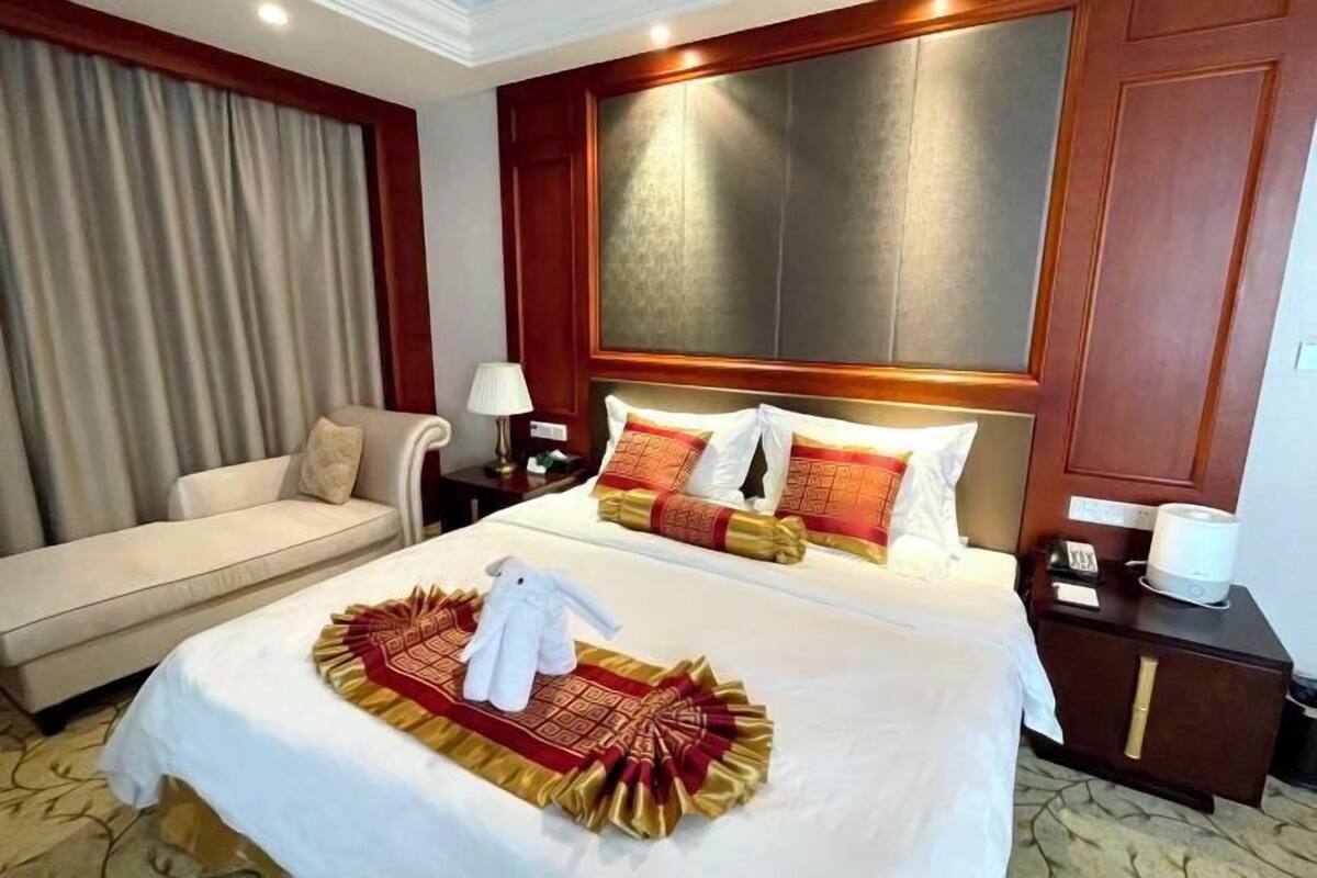 A hotel room with a large bed, a sofa, a nightstand with a lamp, and a large window with curtains.