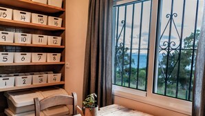 A room with numbered storage bins on shelves, a wooden table, a chair, a metal watering can, and a window with a view of greenery and a fence. - Wild House Tarifa - Hostel (Tarifa)