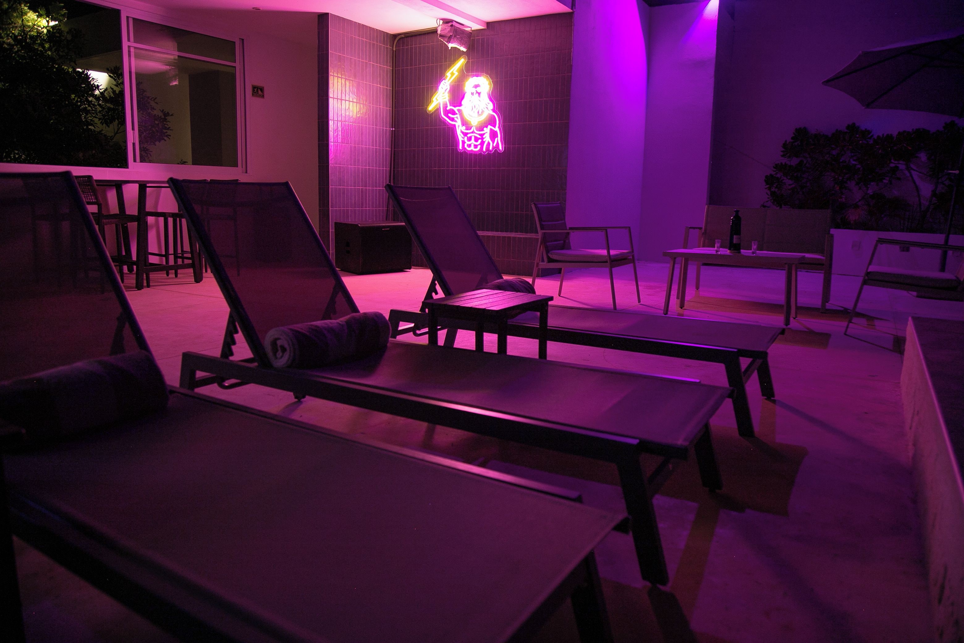 A poolside area with lounge chairs and tables, a neon sign, and a building with a tiled exterior.