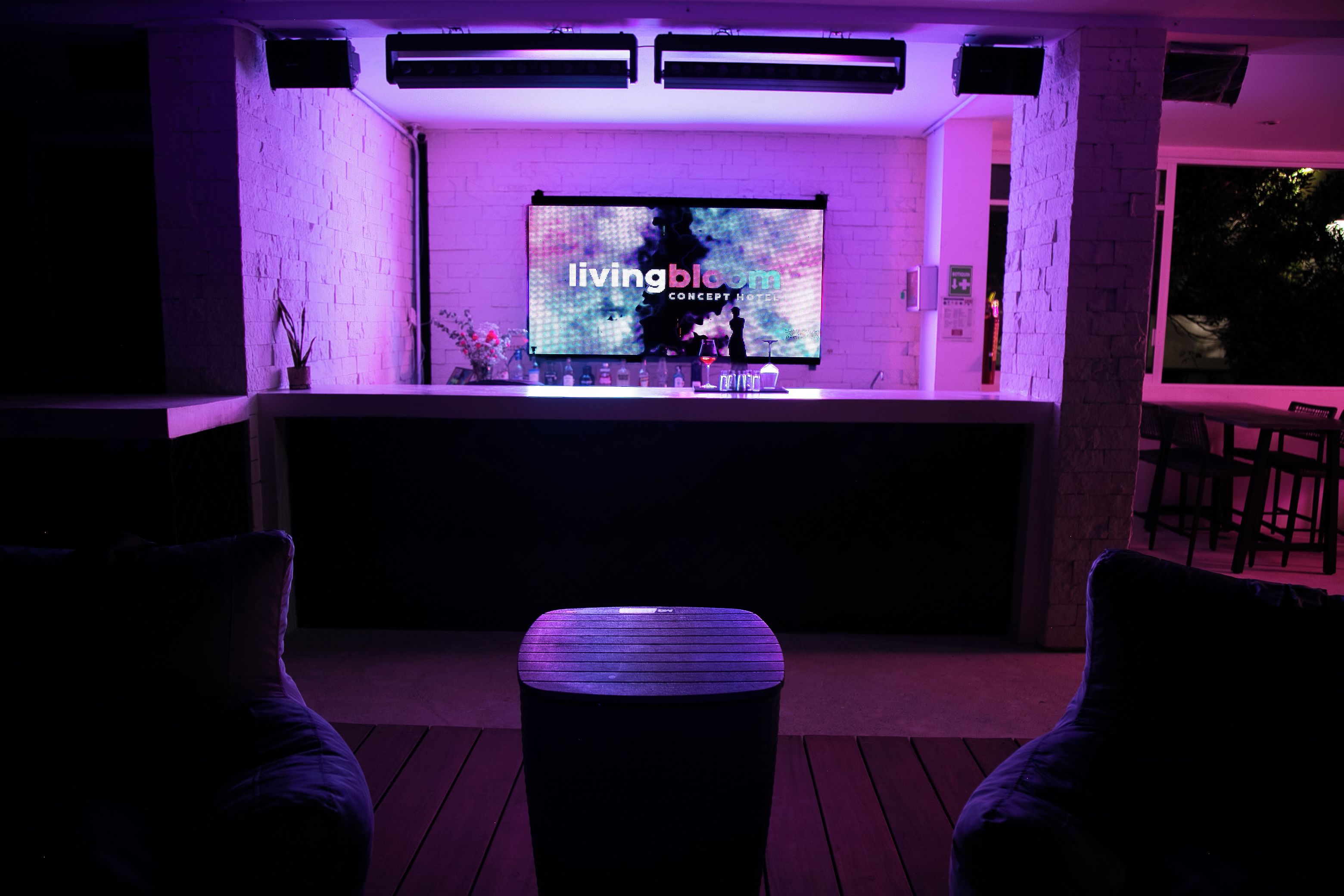A bar area with a flat-screen TV displaying 'livingbloom concept hotel' and a bar counter with bottles and glasses.