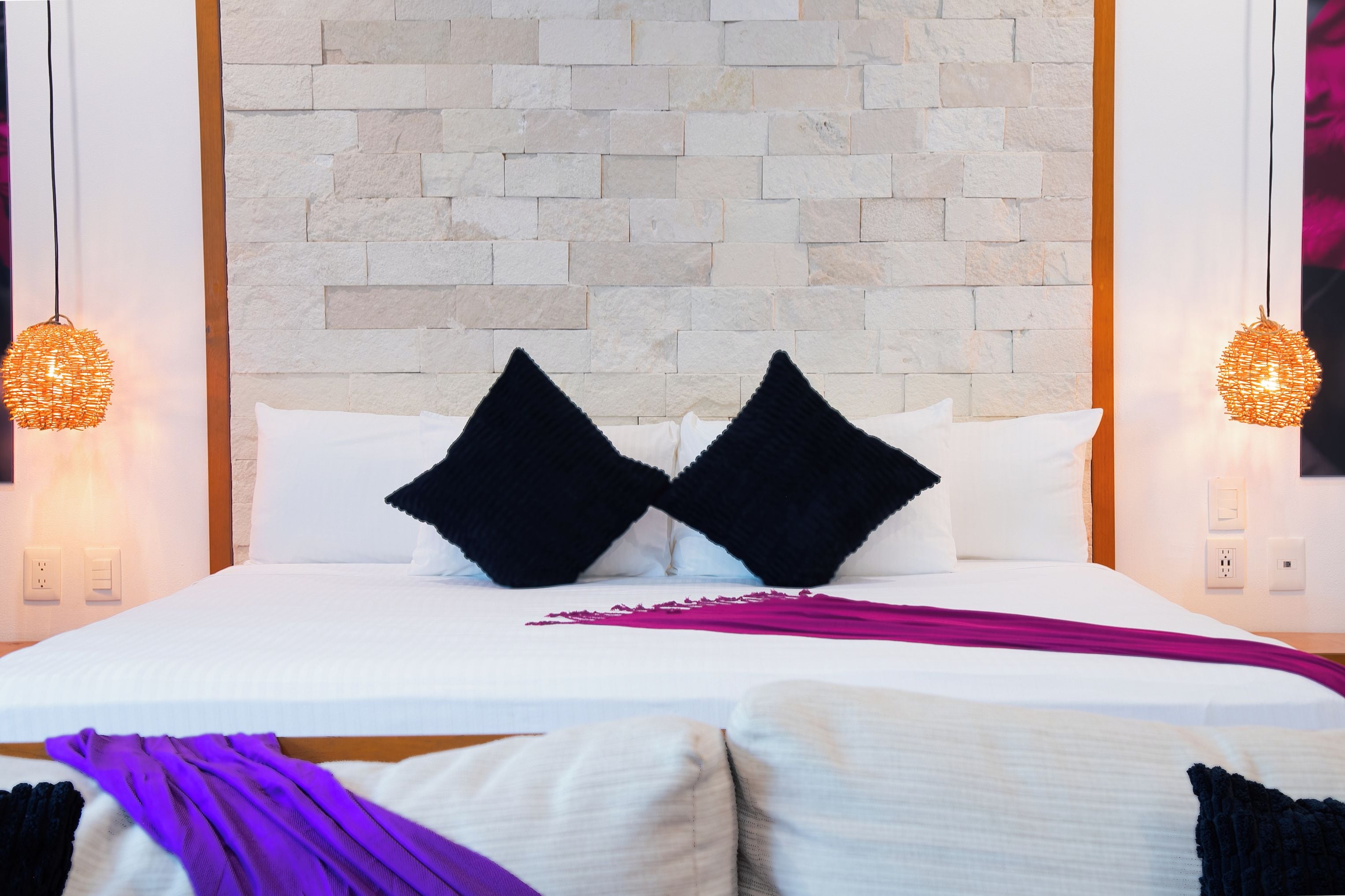 A hotel room with a brick wall, two black pillows, a white bedspread, and two wall-mounted lamps.
