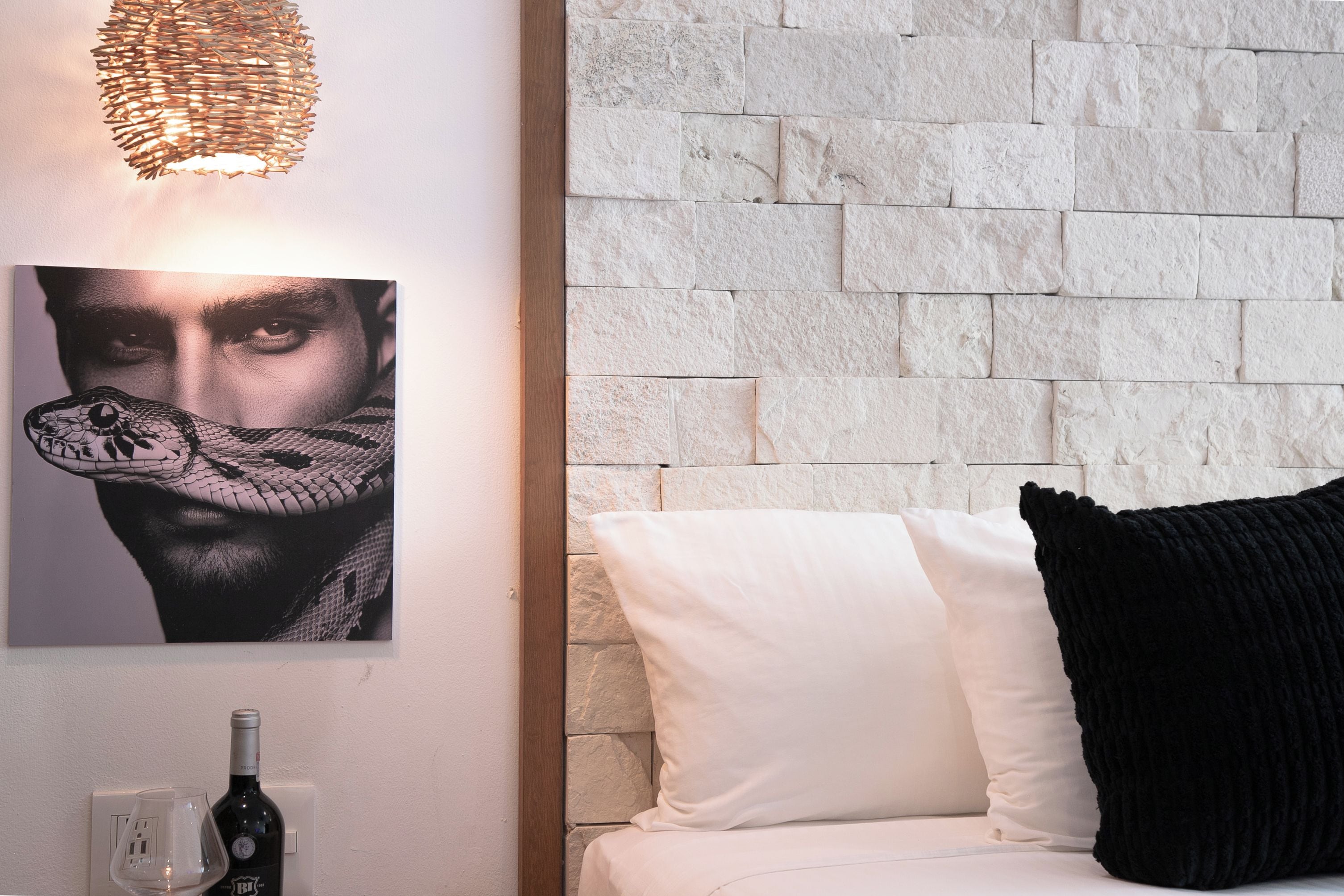 Une pièce avec un mur en briques blanches, un canapé blanc avec un coussin noir, un tableau encadré accroché au mur et une bouteille de vin posée sur le sol.