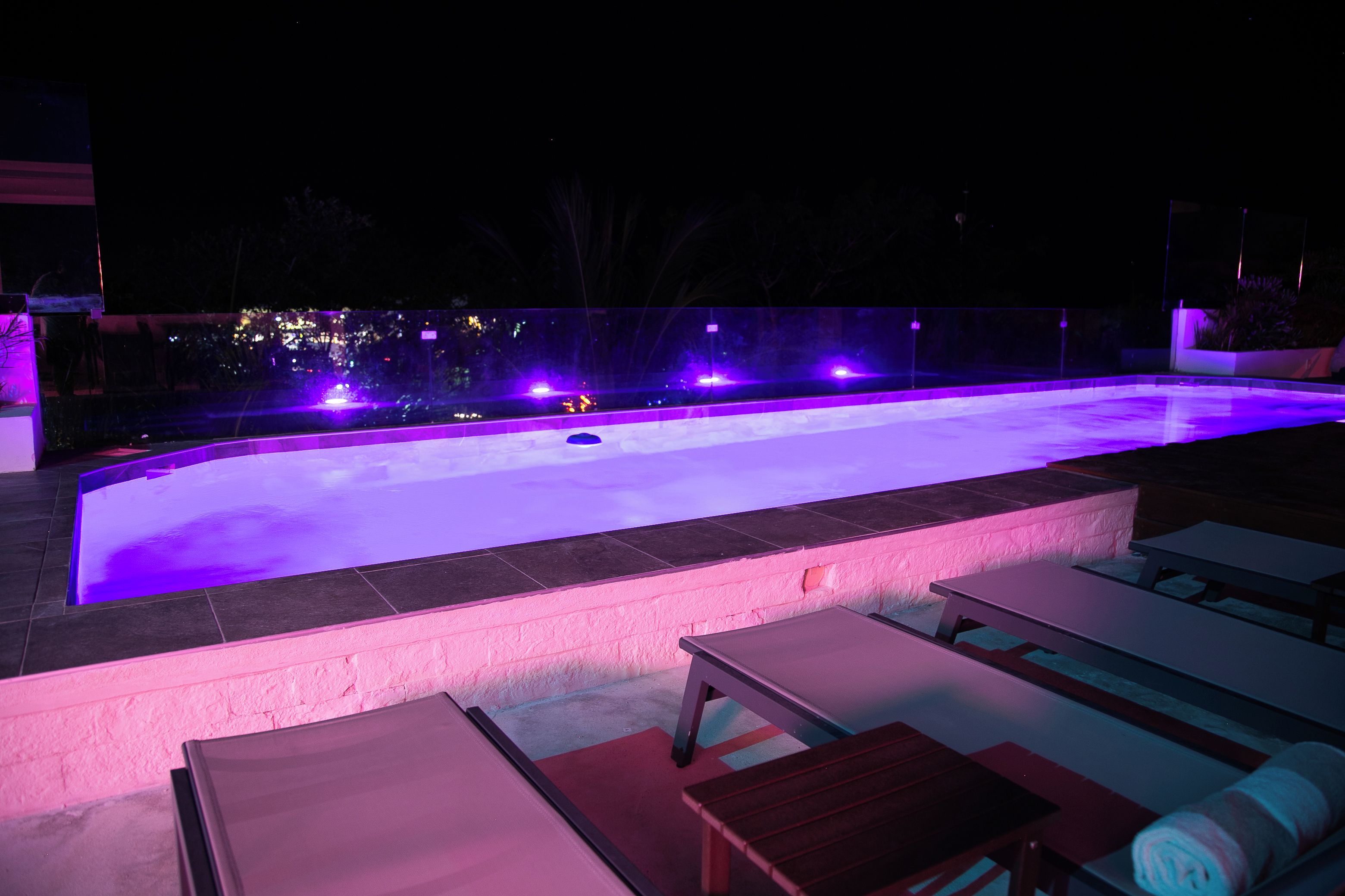Ein Pool mit geschwungener Kante, umgeben von Liegestühlen und einer Bank, mit einer Stadtlandschaft im Hintergrund.