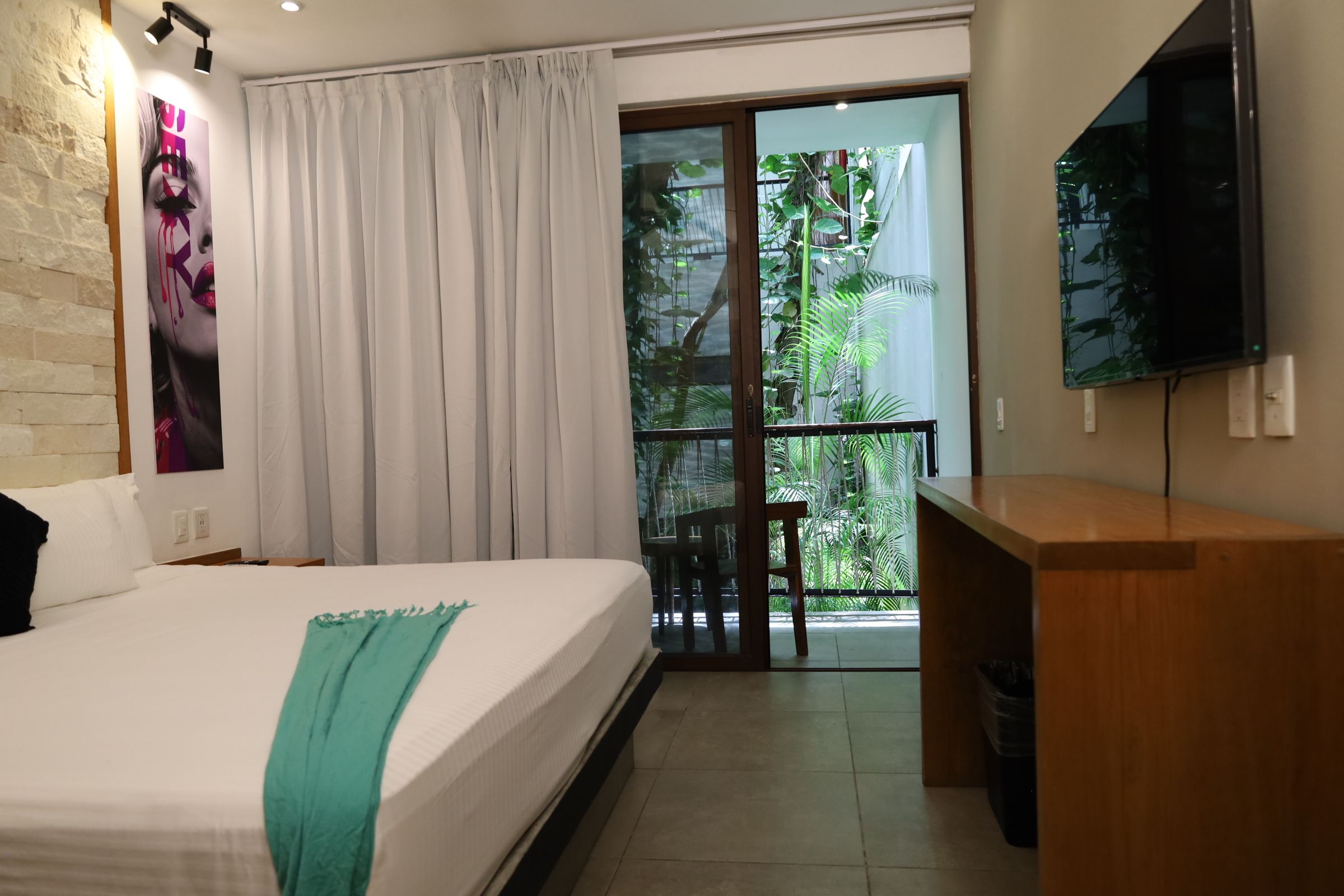 Habitación de hotel con cama, escritorio, televisión y balcón con plantas.