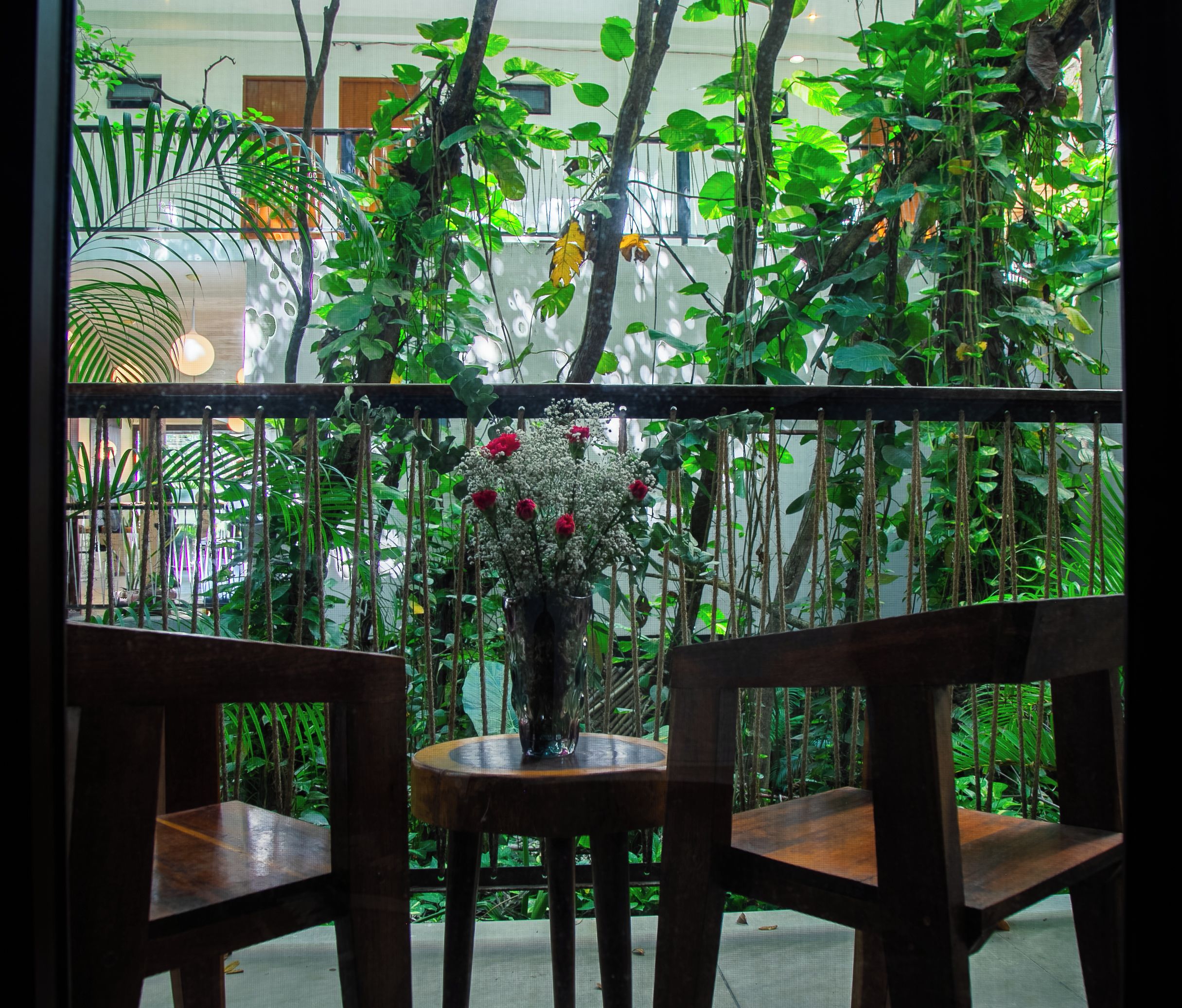 A view through a window with a wooden table and chairs, a vase with flowers, and lush green plants.