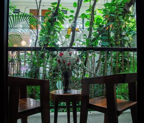 Vista a través de una ventana que muestra una mesa y sillas de madera, un florero con flores y plantas verdes exuberantes.
