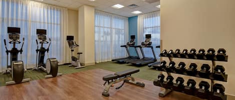 Un gimnasio con cintas de correr, elípticas, una prensa de banca y un estante con mancuernas.