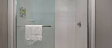 A glass-enclosed shower with a white towel on a metal rack, a showerhead, and a small shelf with toiletries.