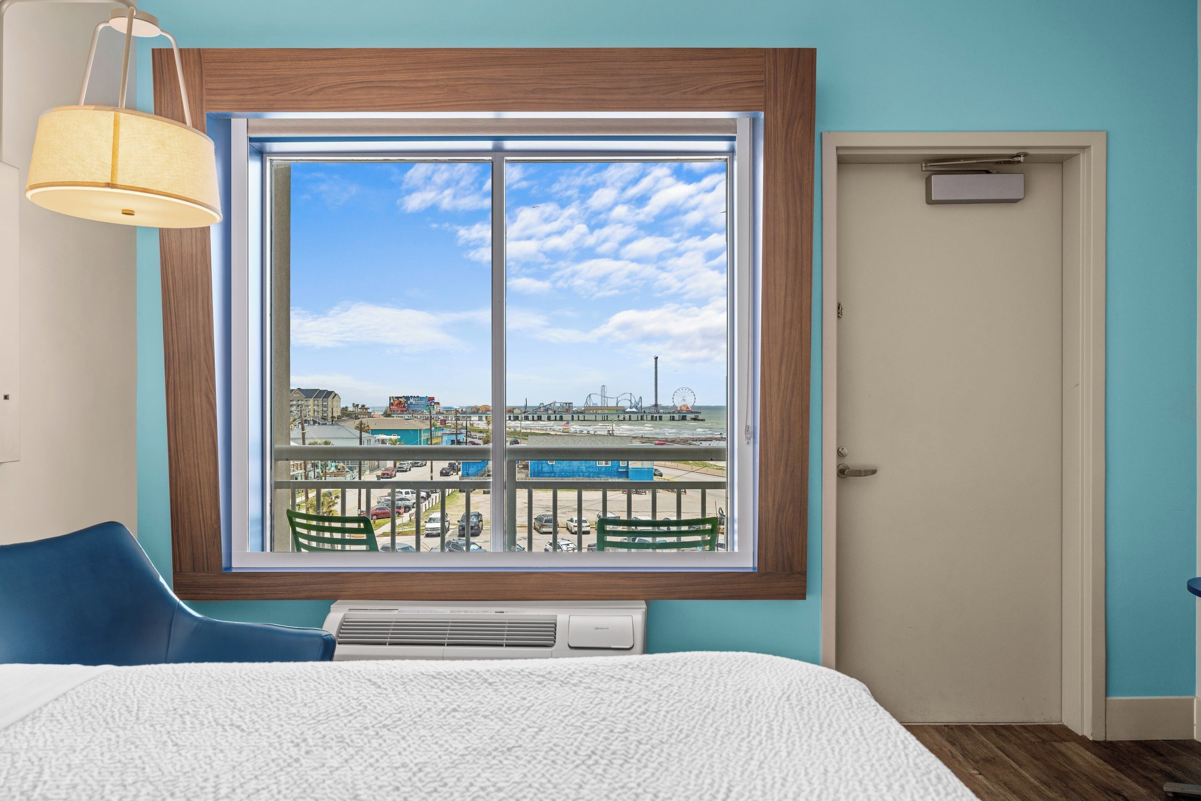 A hotel room with a large window, a bed, a chair, a desk, and a door.