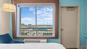 A hotel room with a large window, a bed, a chair, a desk, and a door.