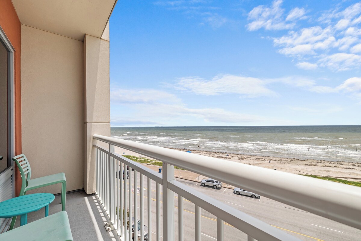 A balcony with a view of the beach and ocean, a road with cars, and a railing.