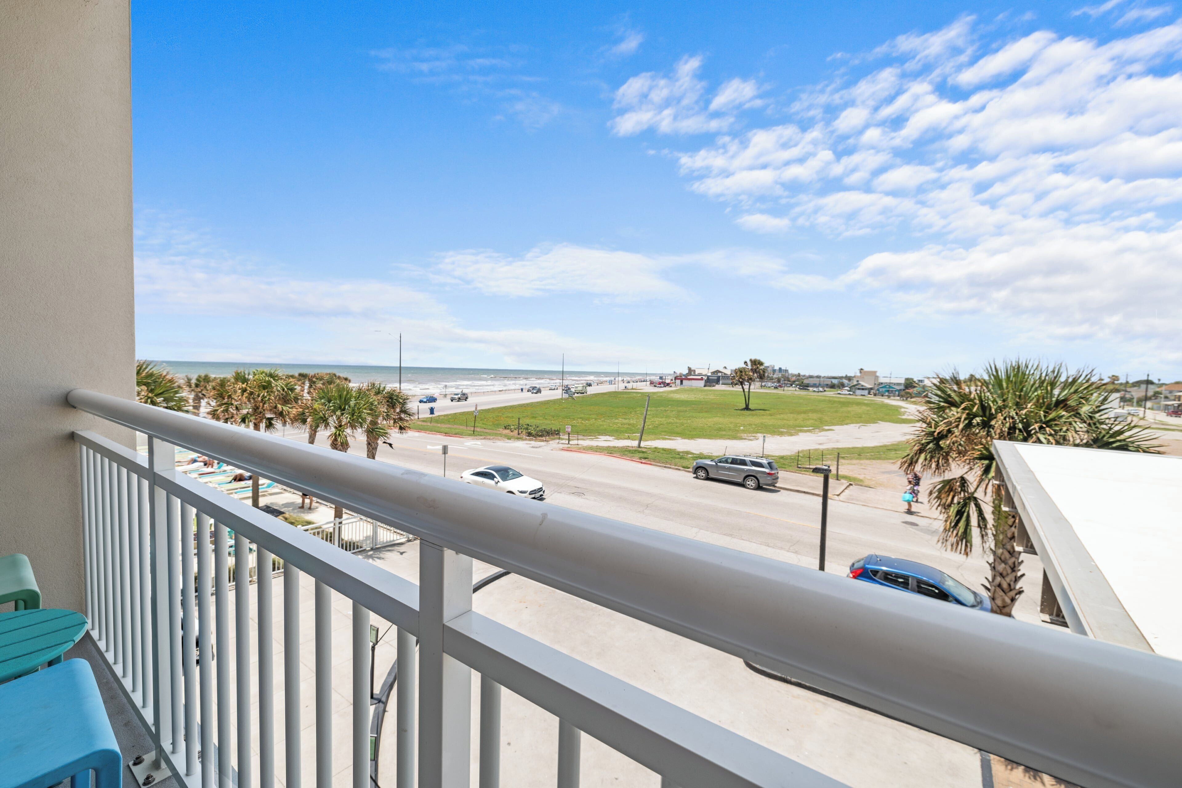 A balcony with a view of a beach, a road with cars, and a grassy area with palm trees.