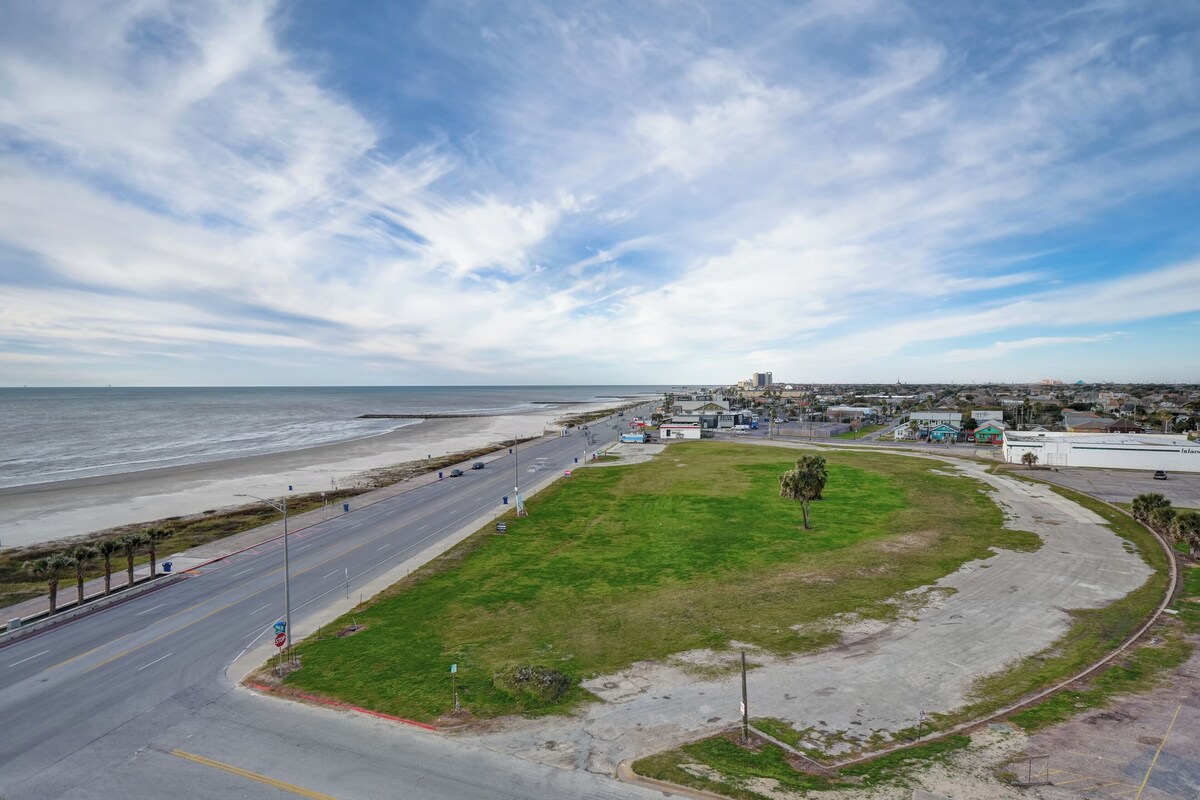 A coastal road with a grassy area, a beach, and buildings in the distance.