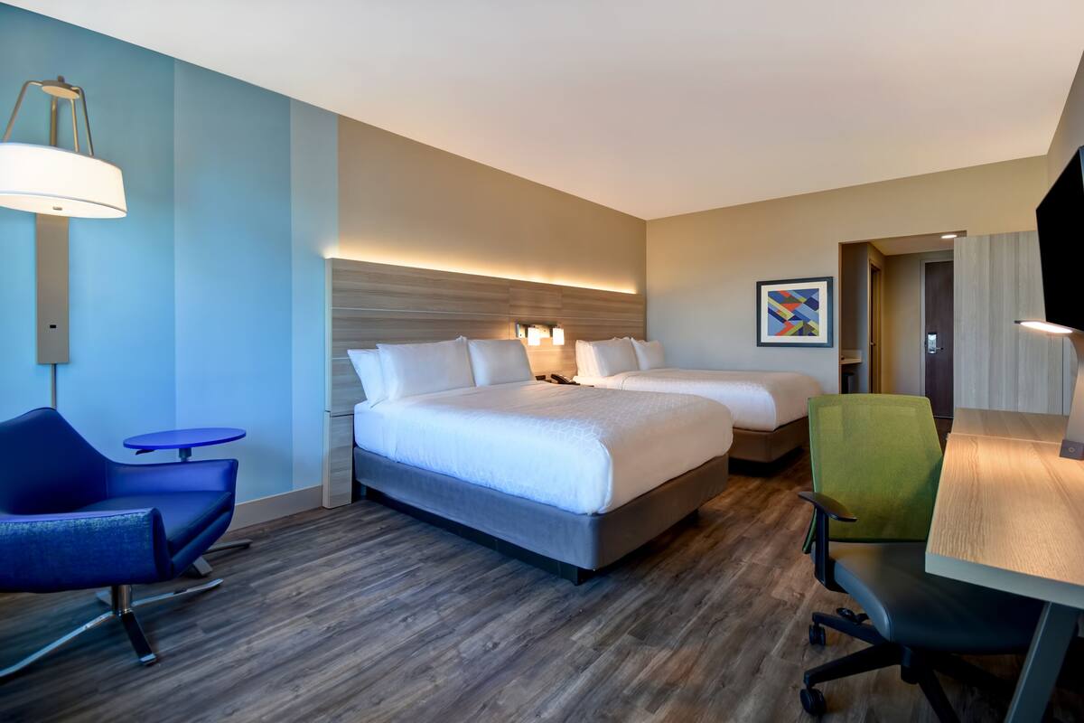 A hotel room with two beds, a desk, a chair, a lamp, and a painting on the wall.