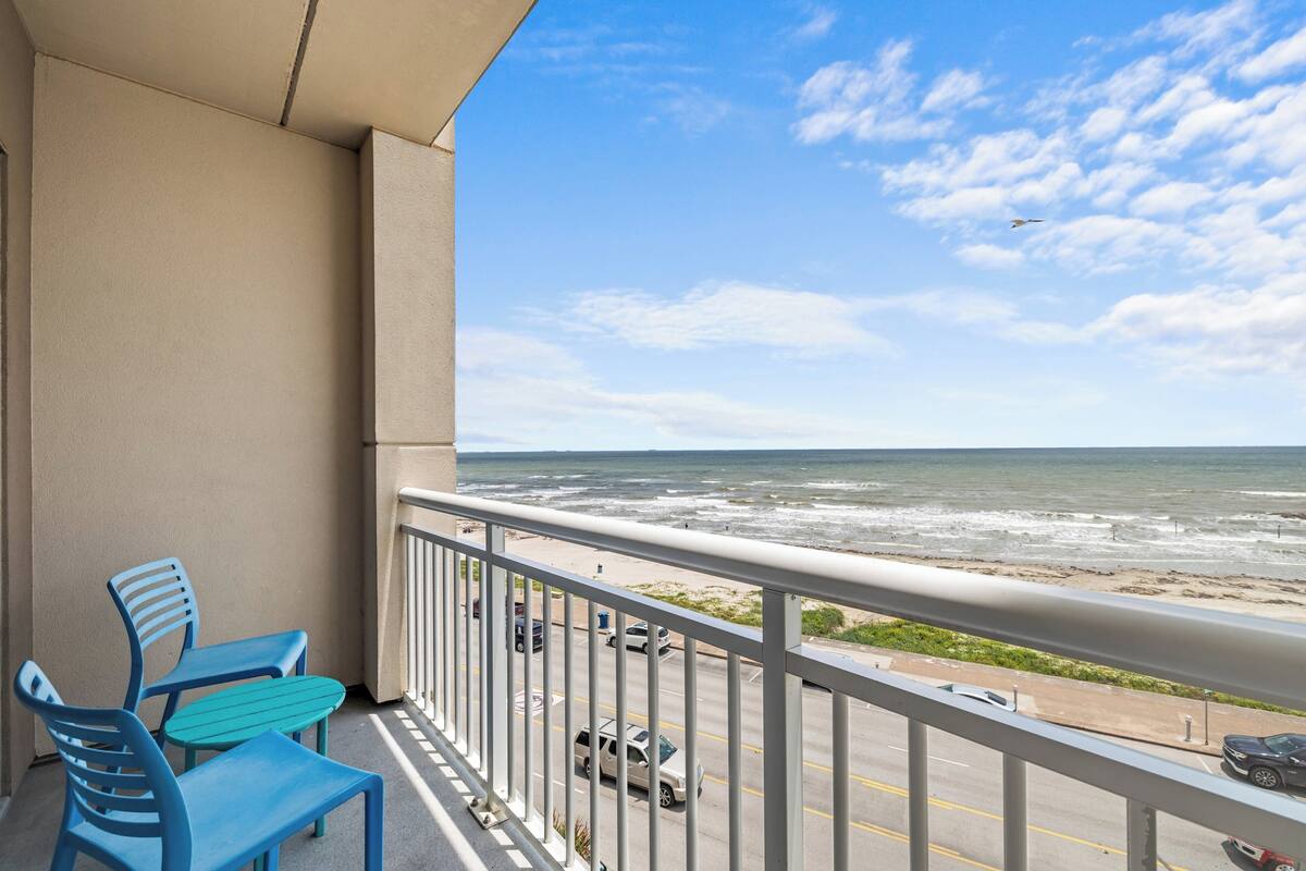 A balcony with two blue chairs, a view of the beach, and a railing.