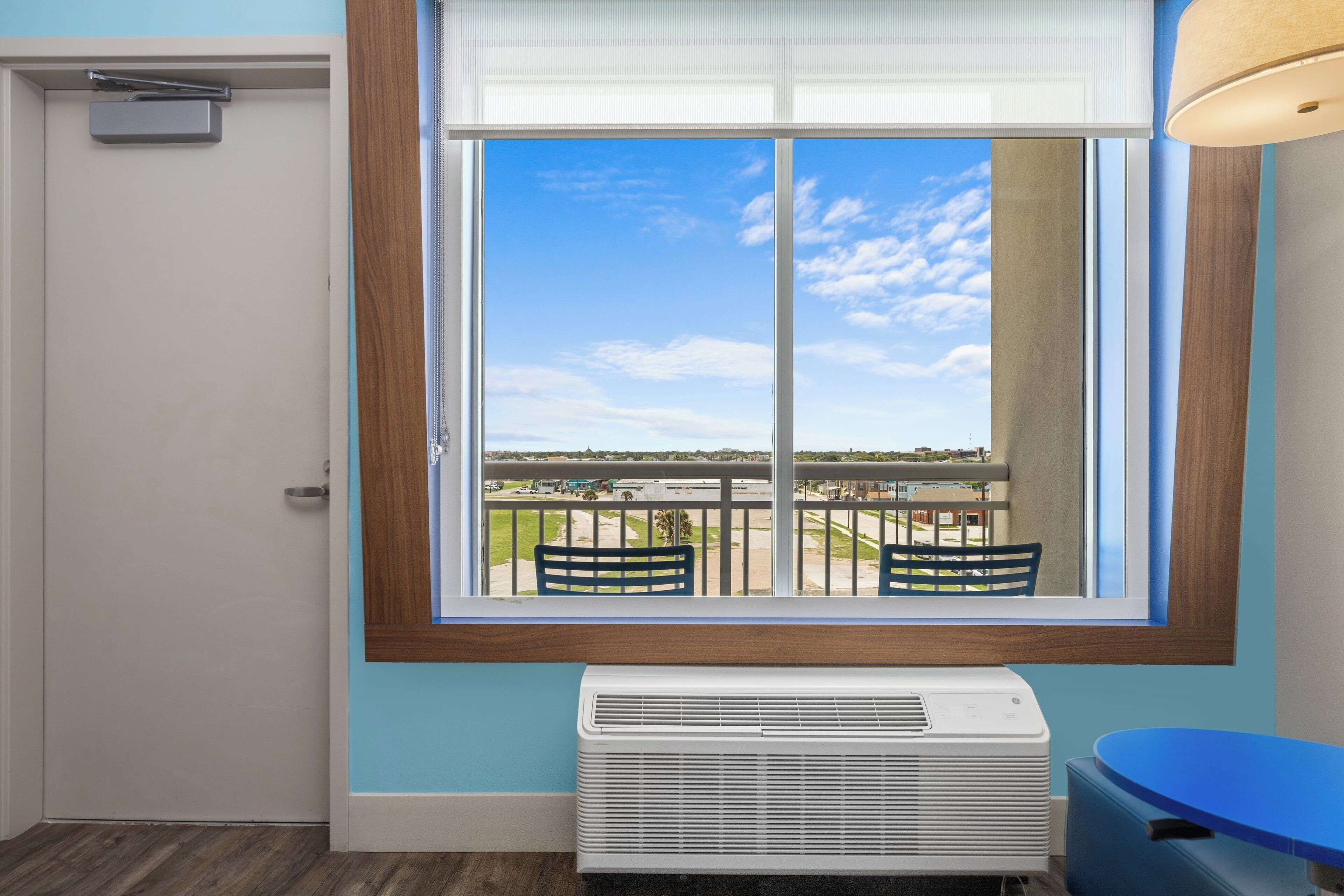 A room with a large window, a white air conditioning unit, a blue table, and a door.
