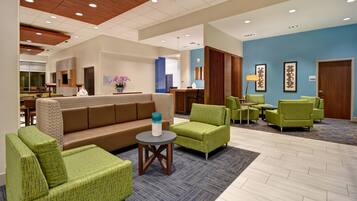Un hall de hotel con un sofá marrón, sillones verdes, una mesa redonda pequeña y un jarrón azul.