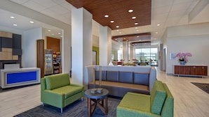 A modern lobby with green upholstered sofas, a reception desk, a refrigerator stocked with snacks, and a large window offering a view of the city. - Holiday Inn Express & Suites Galveston Beach by IHG (Galveston)