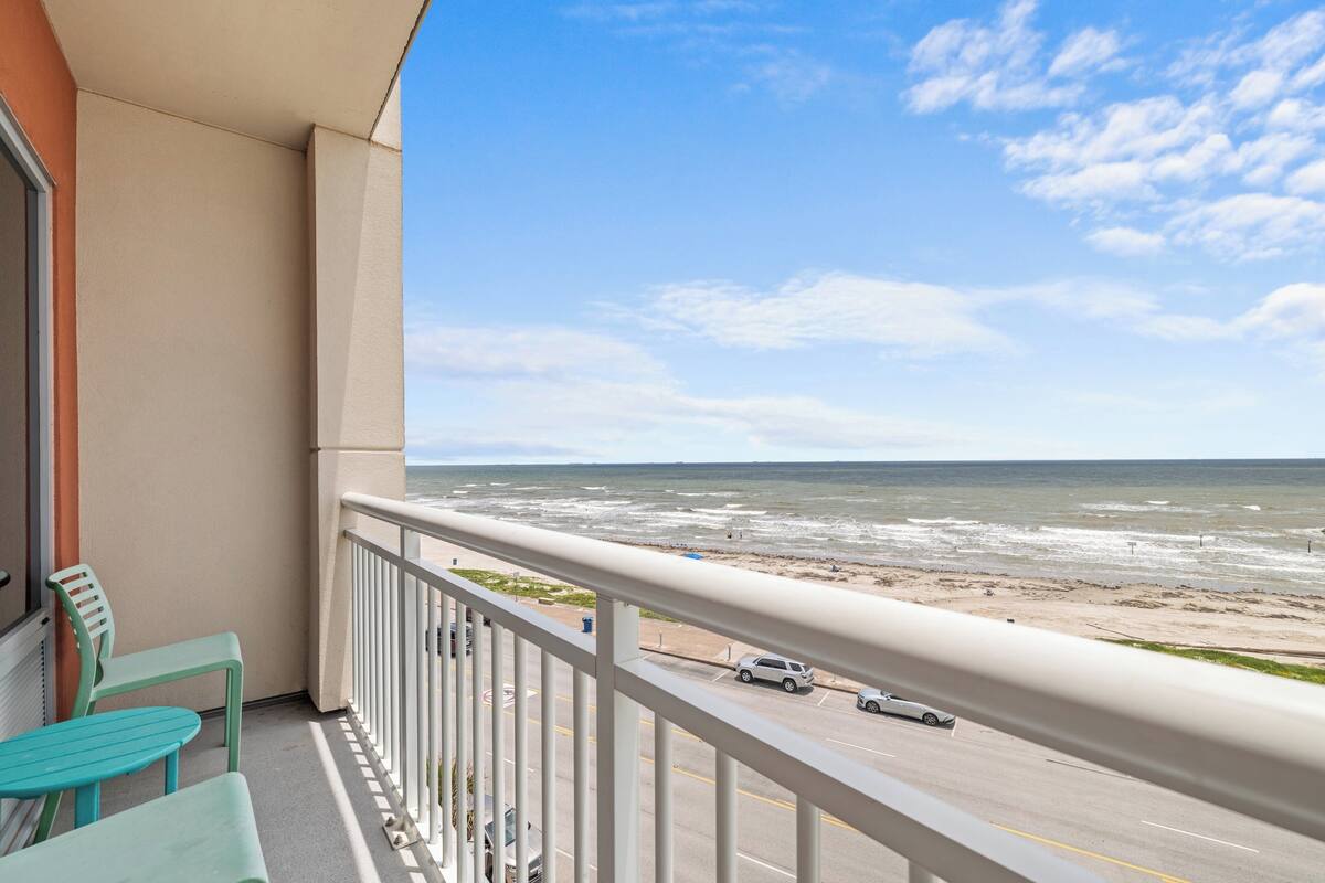 A balcony with a view of the beach and ocean, a road with cars, and a railing.