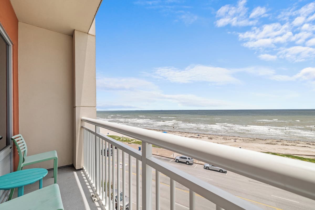 A balcony with a view of the beach and ocean, a road with cars, and a railing.