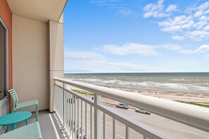 Un balcon avec vue sur la plage et l’océan, une route où circulent des voitures et une rambarde.