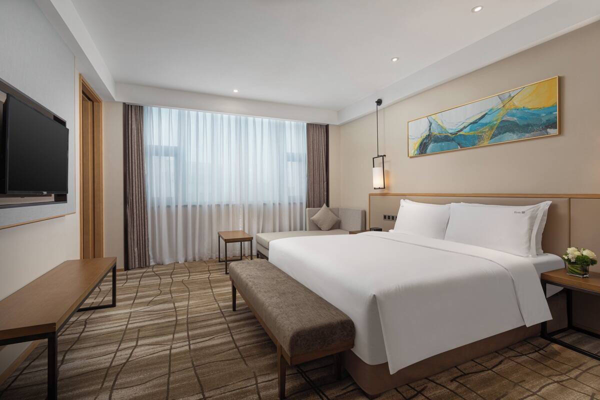 A hotel room with a large bed, a small bedside table, a bench, a TV, and a painting on the wall.