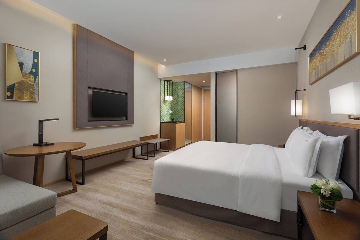 A modern hotel room with a large bed, a flat-screen TV, a small desk, and a bench.
