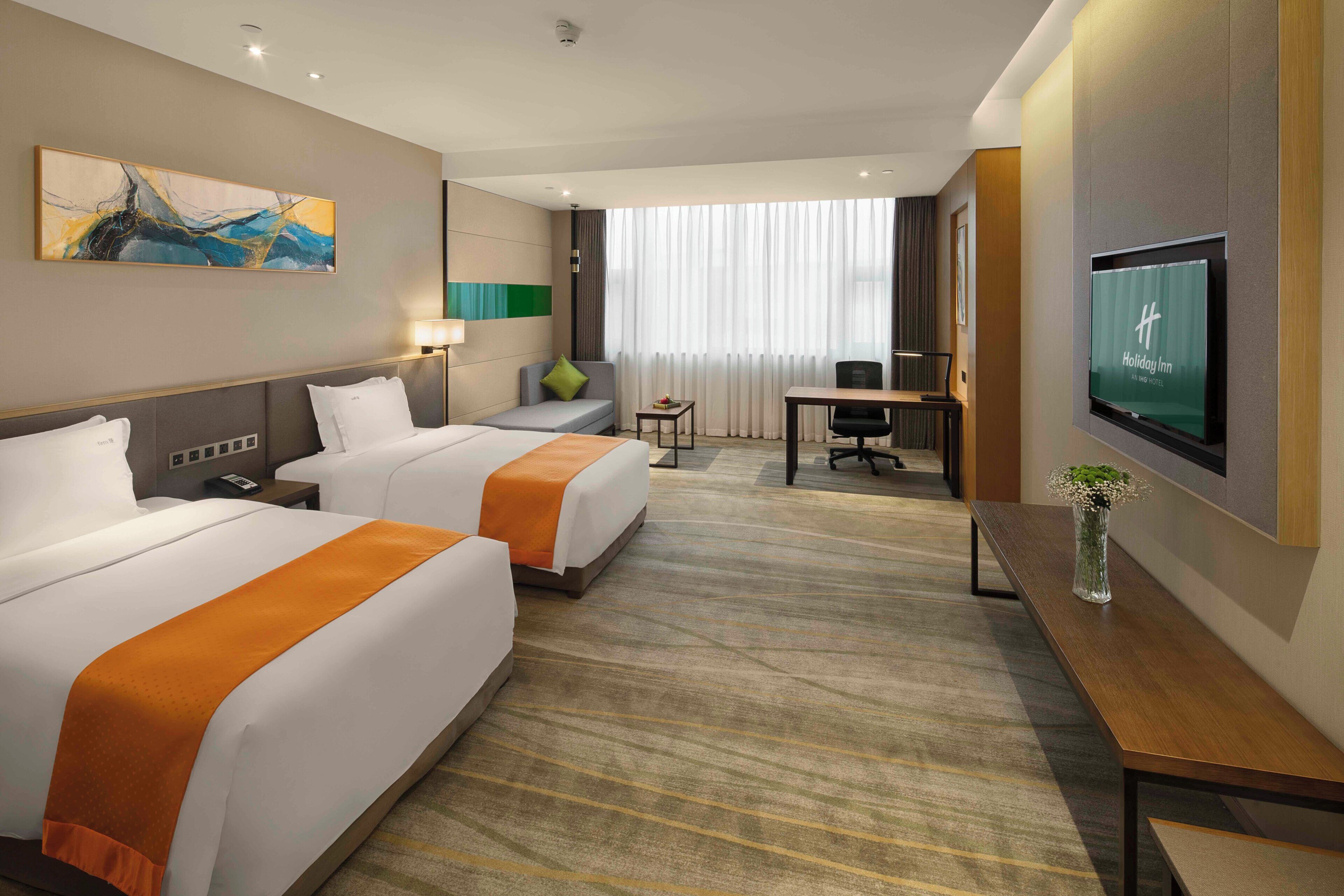 A hotel room with two beds, a desk, a TV, and a large window with curtains.