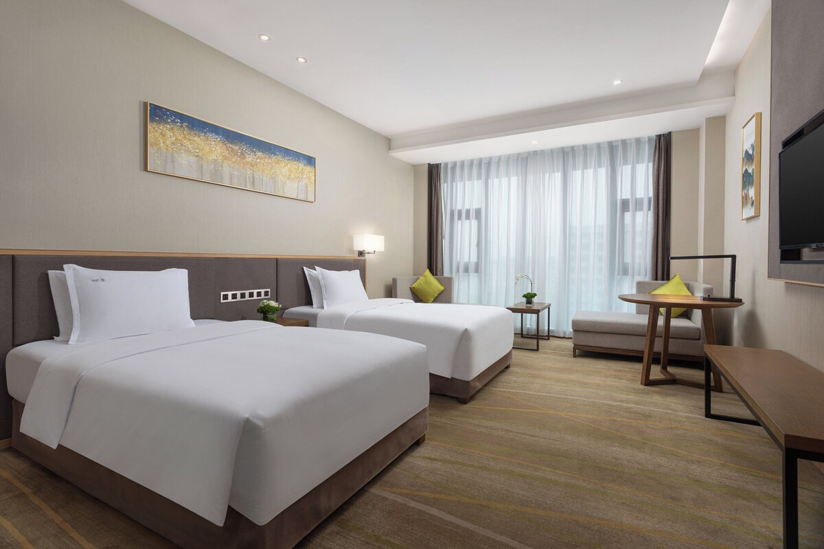 A hotel room with two beds, a desk, a chair, a TV, and a large window with curtains.