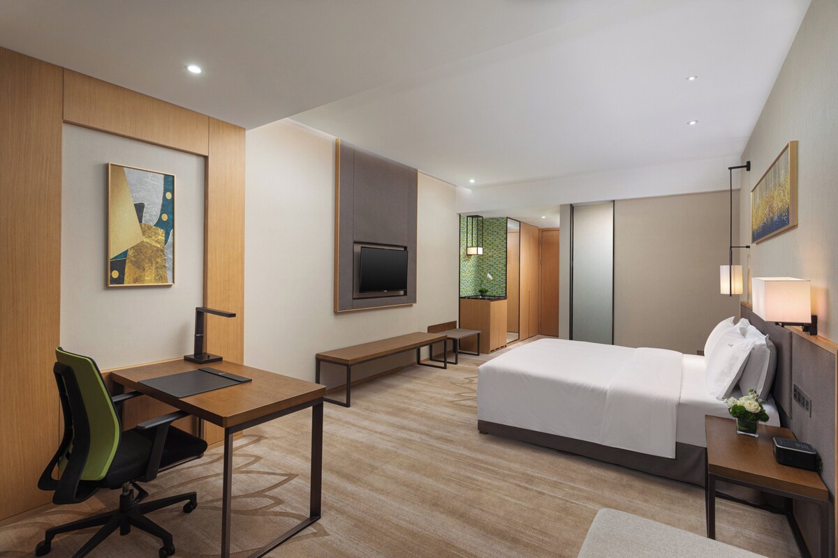 A modern hotel room with a large bed, a desk with a chair, a television, and a small bench.