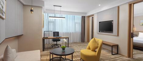 A hotel room with a sofa, a yellow armchair, a round table, a TV, a desk with a chair, and a large window with curtains.