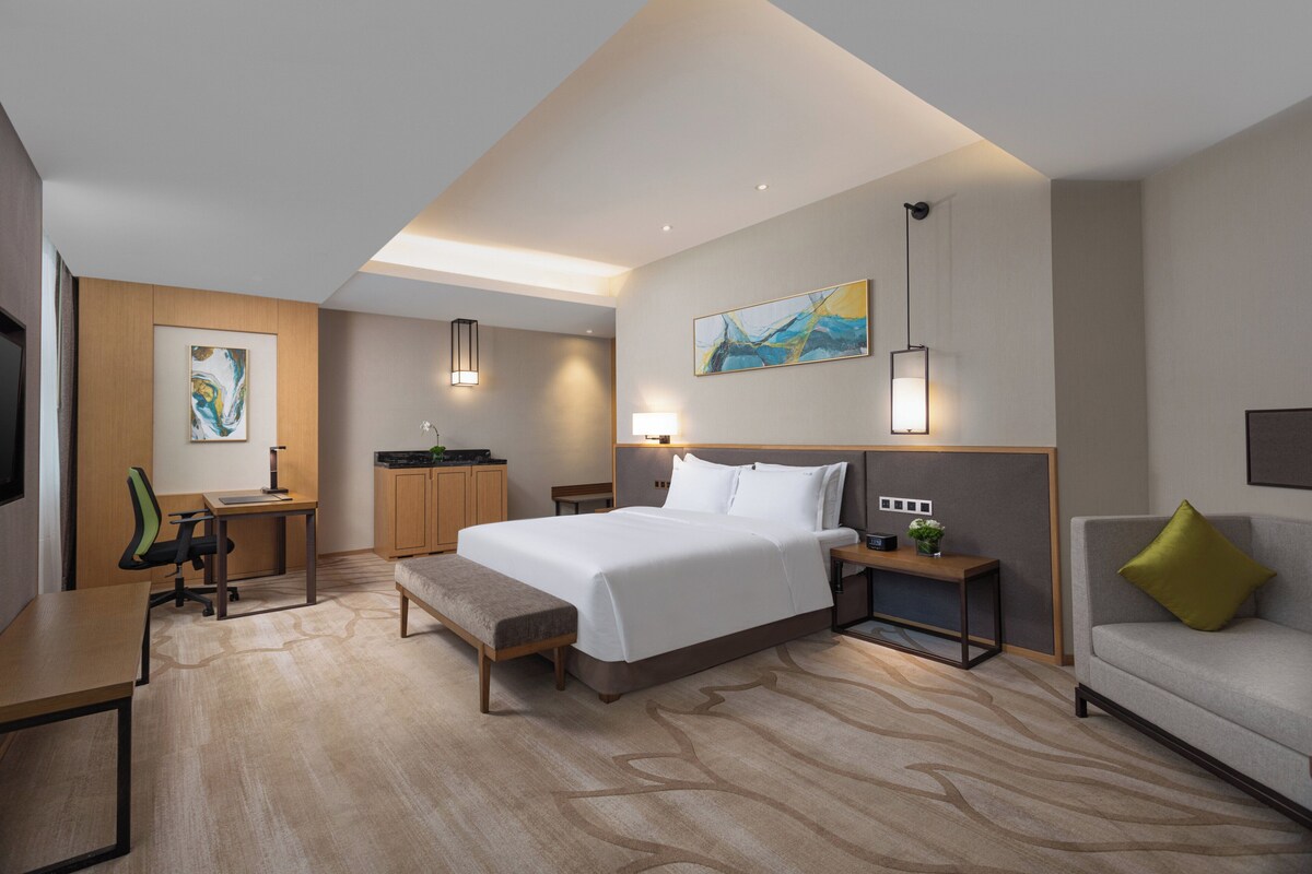 A hotel room with a bed, desk, chair, bedside table, lamp, and a sofa.