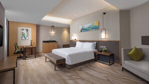 A hotel room with a bed, desk, chair, bedside table, lamp, artwork, and a sofa.