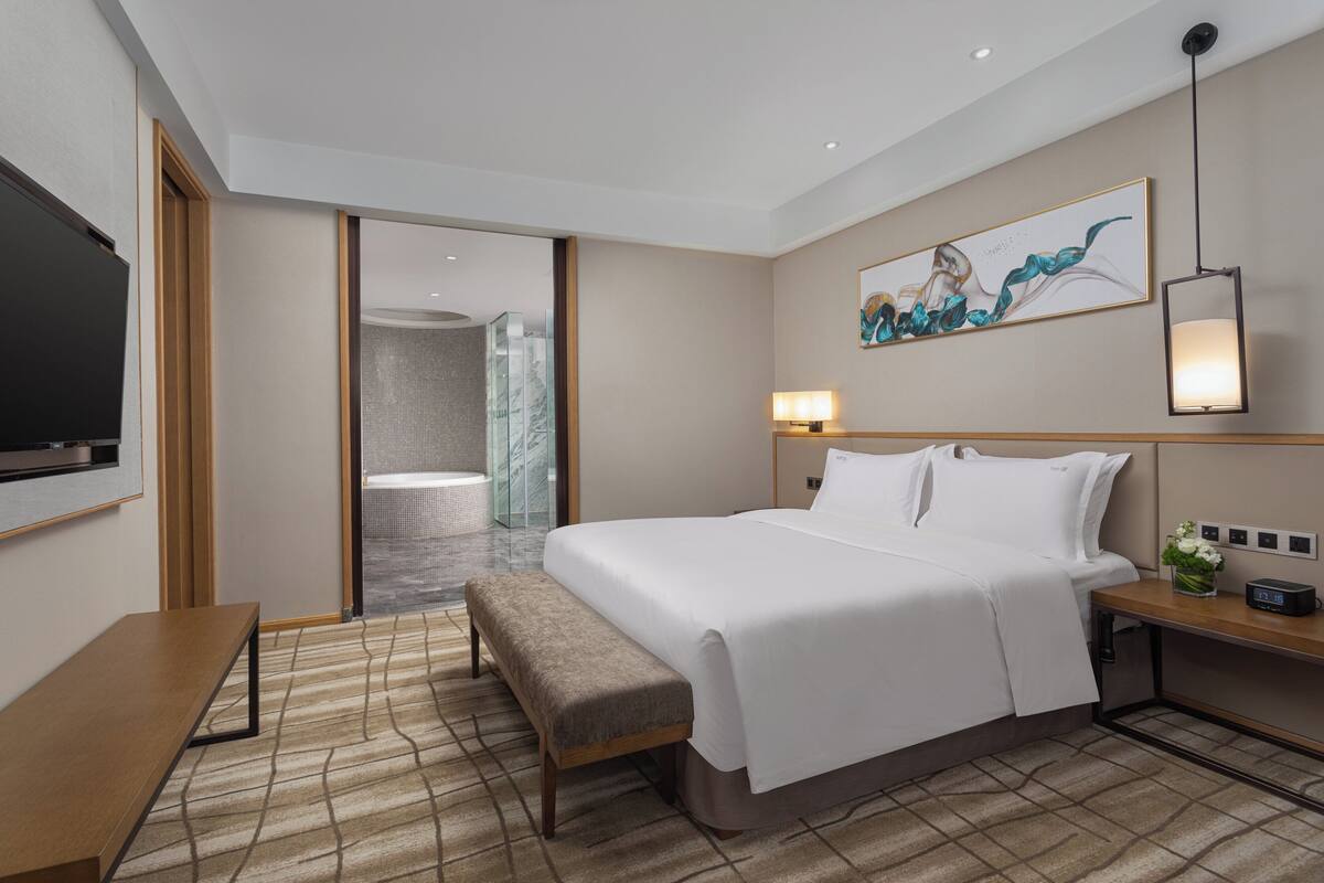 A modern hotel room with a large bed, bedside table, bench, and a TV mounted on the wall.