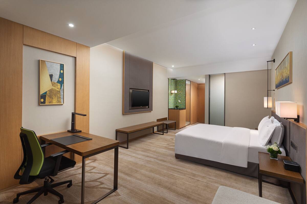 A modern hotel room with a large bed, a desk with a chair, a television, and a small bench.