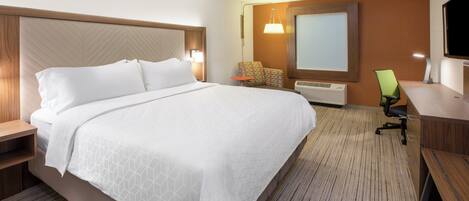 A hotel room with a large bed, bedside tables, a desk with a chair, a television, and an air conditioning unit.
