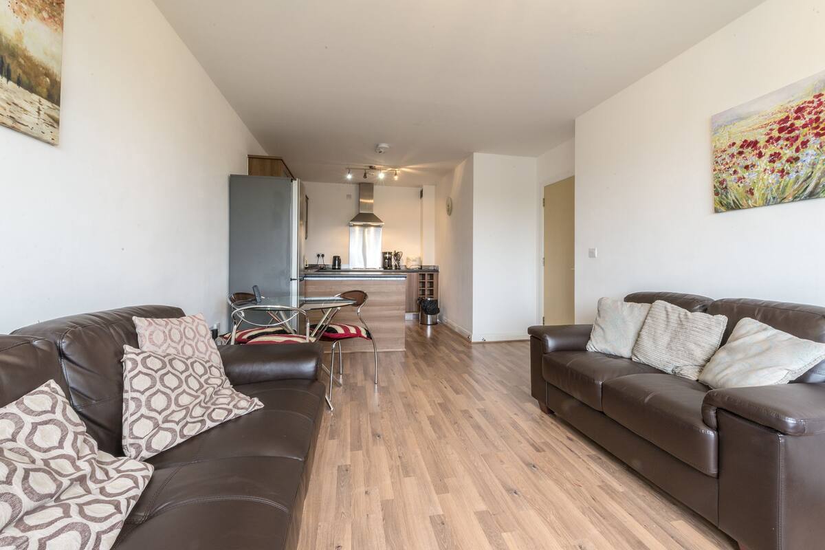 A modern living room with two leather sofas, patterned cushions, a dining table with chairs, and a kitchen in the background.