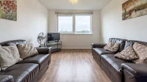Un salon avec deux canapés en cuir, un téléviseur posé sur un meuble, un ventilateur et une grande fenêtre donnant sur des immeubles.