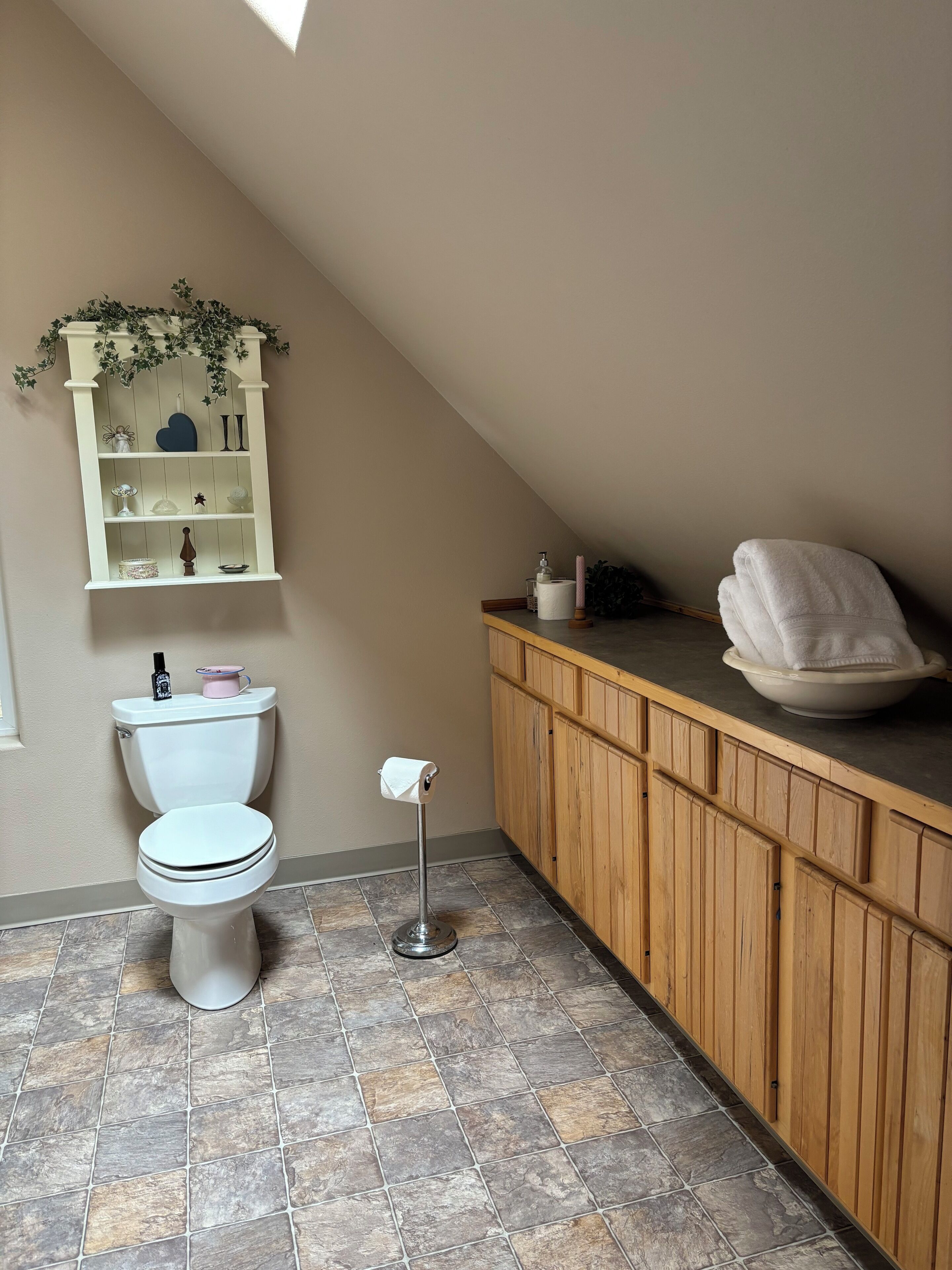Un baño con inodoro, tocador de madera, estante con adornos y piso de baldosas.