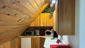 Cocina compacta con paredes y techo de madera, un refrigerador blanco, un microondas, una cafetera y un lavabo con un felpudo rojo.