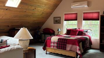Ein gemütliches Schlafzimmer im Dachgeschoss mit schrägen Wänden, einem Deckenventilator, einem Bett mit rot-schwarzer Karomusterdecke, einer Lampe und einem Fenster mit roten Rollläden.