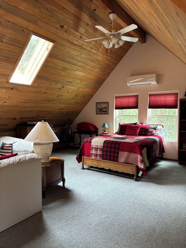 A cozy attic bedroom with a sloped ceiling, a ceiling fan, a bed with a red and black checkered bedspread, a lamp, and a window with red blinds.