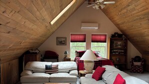 Un dormitorio acogedor en el ático, con techo inclinado, ventilador de techo, dos ventanas con cortinas rojas, una mesa de madera, un sofá y una lámpara.